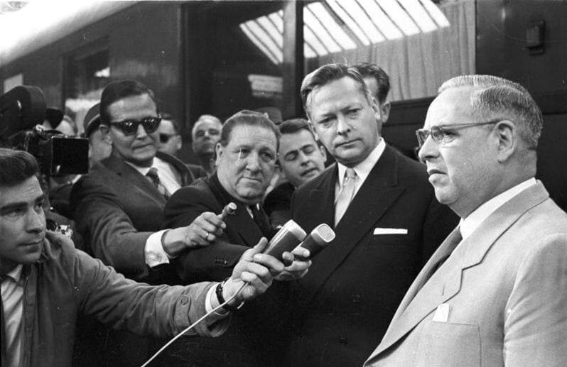 El periodista Karl-Eduard von Schnitzler con gafas de sol sostiene un micrófono entre la prensa en la Conferencia de Ministros de Ginebra.