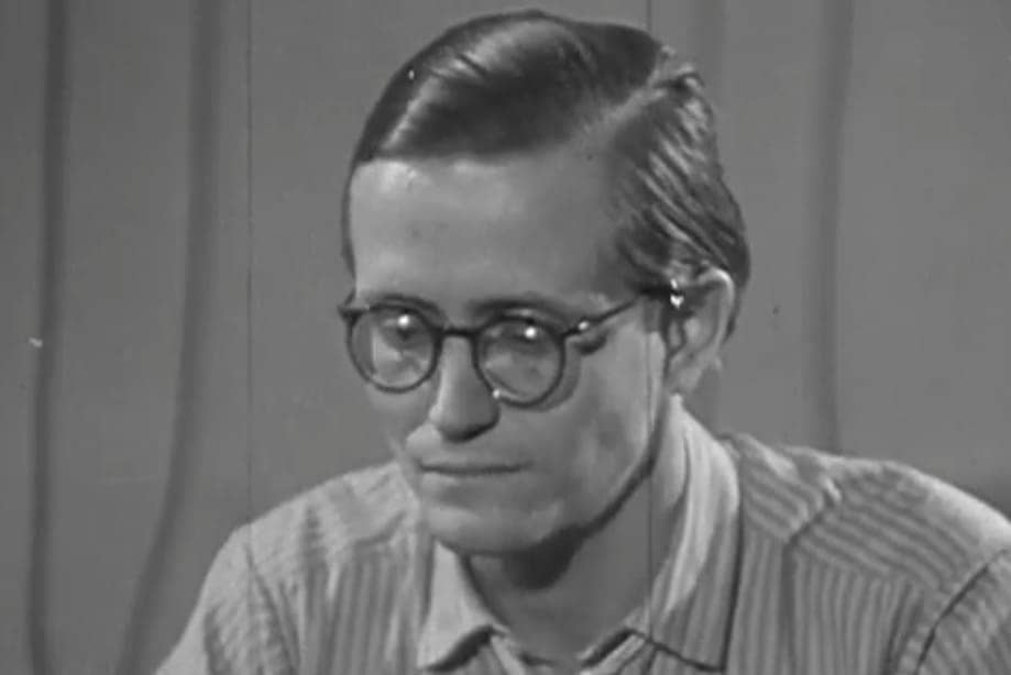 Primer plano de Karl-Eduard von Schnitzler con gafas redondas de metal y camisa de rayas mirando fijamente durante una transmisión televisiva.