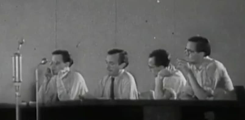 Cuatro hombres de camisa clara sentados en una mesa larga de conferencias con micrófonos, con Schnitzler gesticulando en el extremo derecho.