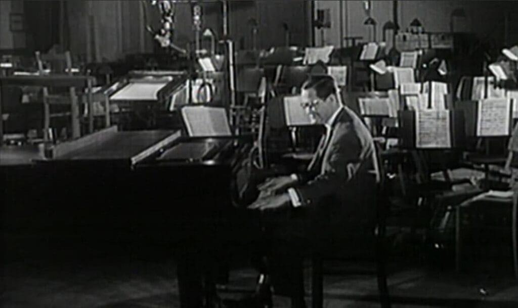 Karl-Eduard von Schnitzler de traje toca un piano de cola en un amplio estudio de grabación musical rodeado de atriles vacíos al fondo.