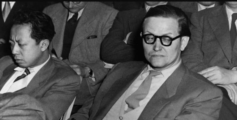 Karl-Eduard von Schnitzler con gafas de pasta mira de reojo sentado junto a un delegado asiático de traje en un congreso internacional.