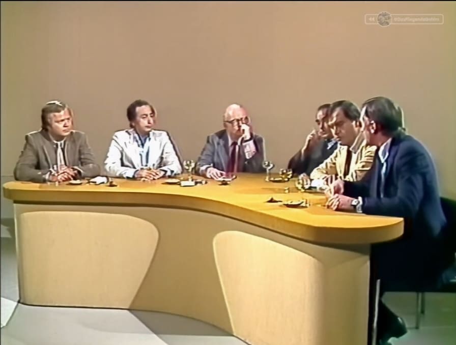 Seis hombres sentados debaten alrededor de una gran mesa curvada de madera en un set de televisión con fondo beige liso.