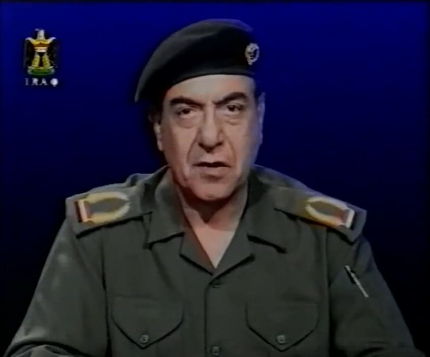 Captura de televisión de Mohamed Said al-Sahaf con uniforme verde y boina negra hablando frente a un fondo azul oscuro.