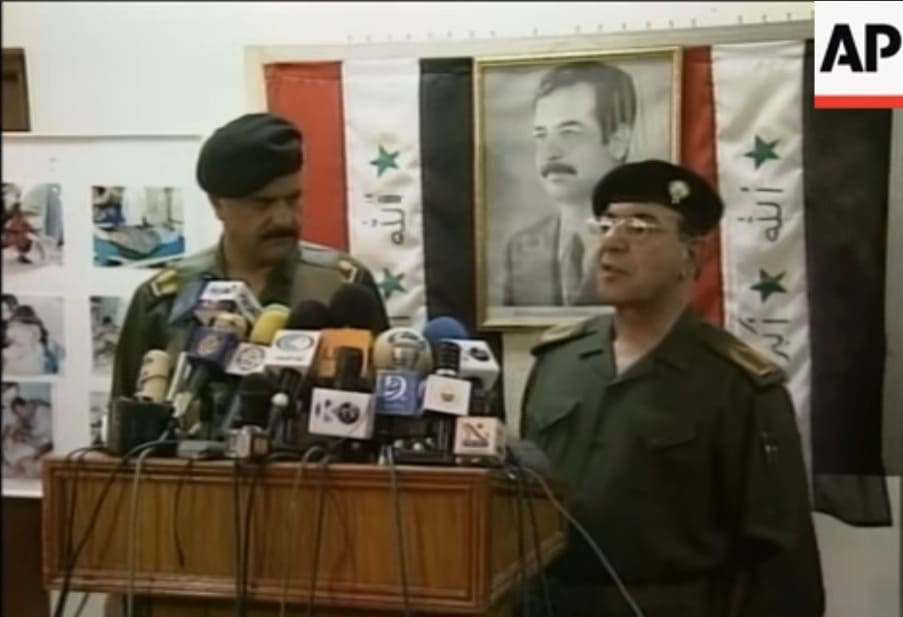 Al-Sahaf da un discurso junto a otro oficial militar frente a banderas de Irak y un gran retrato enmarcado de Saddam Hussein.