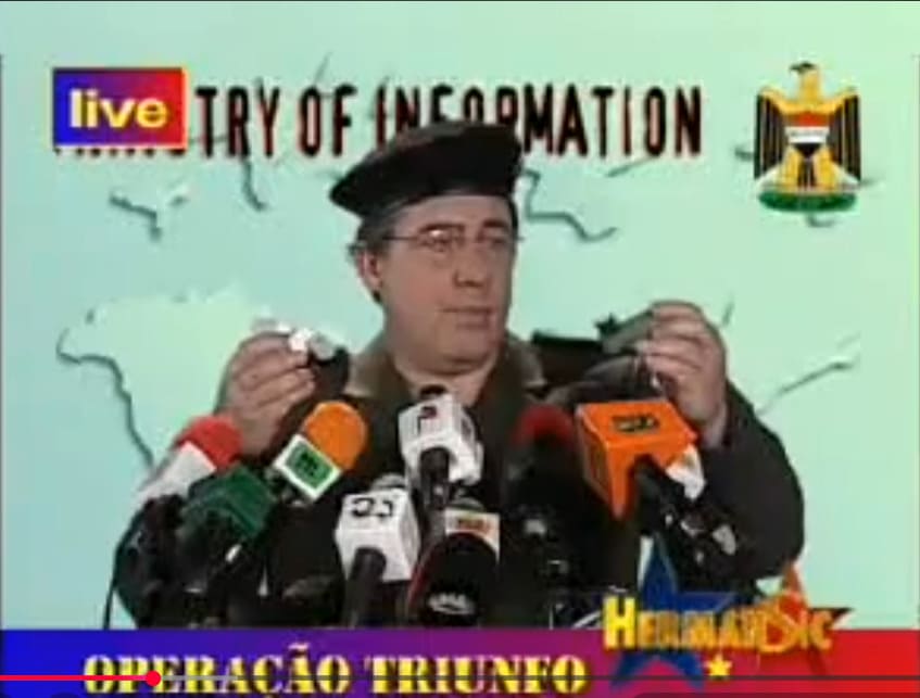 Actor caracterizado como Al-Sahaf sosteniendo dos pequeños objetos metálicos frente a micrófonos en una parodia de televisión.
