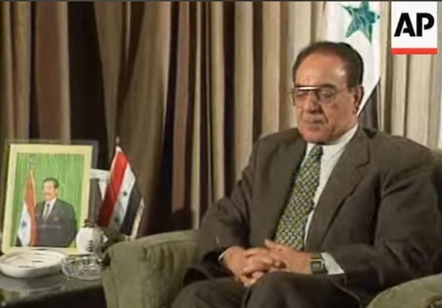 Mohamed Said al-Sahaf en traje gris y gafas sentado en un sillón junto a una bandera iraquí y un retrato de Saddam Hussein.