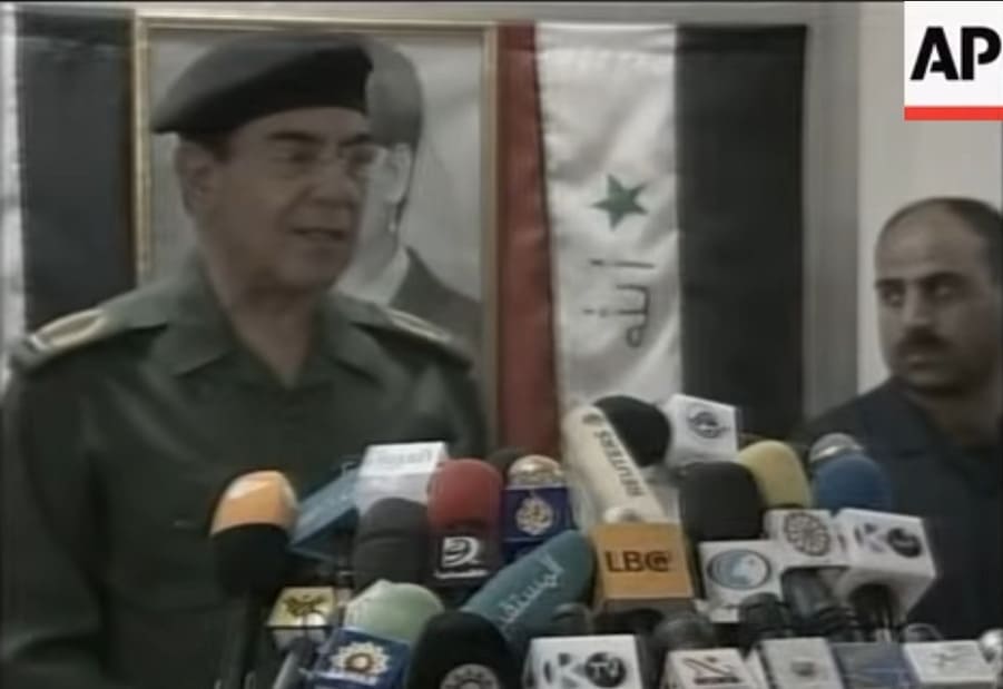 Muhammad Saeed al-Sahhaf con uniforme militar habla ante micrófonos de prensa frente a una foto de Sadam Husein y bandera de Irak.