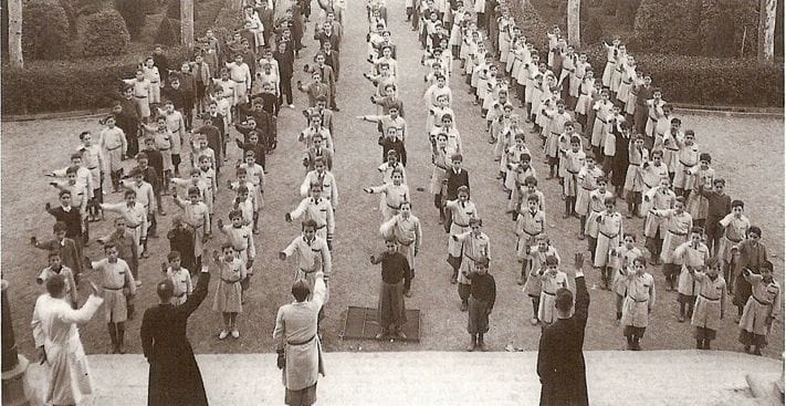 Fotografía en blanco y negro de niños e instructores formados en filas haciendo el saludo fascista al aire libre en un colegio.