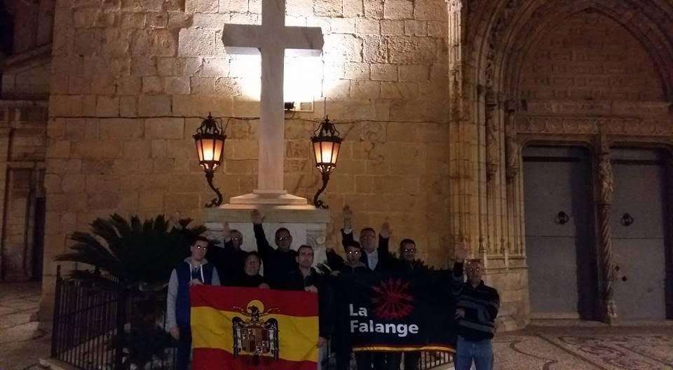Grupo de personas con banderas de Falange y de España haciendo el saludo fascista nocturno frente a una cruz de piedra.