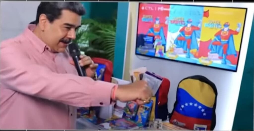 Nicolás Maduro en un evento televisado sosteniendo un micrófono frente a un televisor y morrales de Súper Bigote.