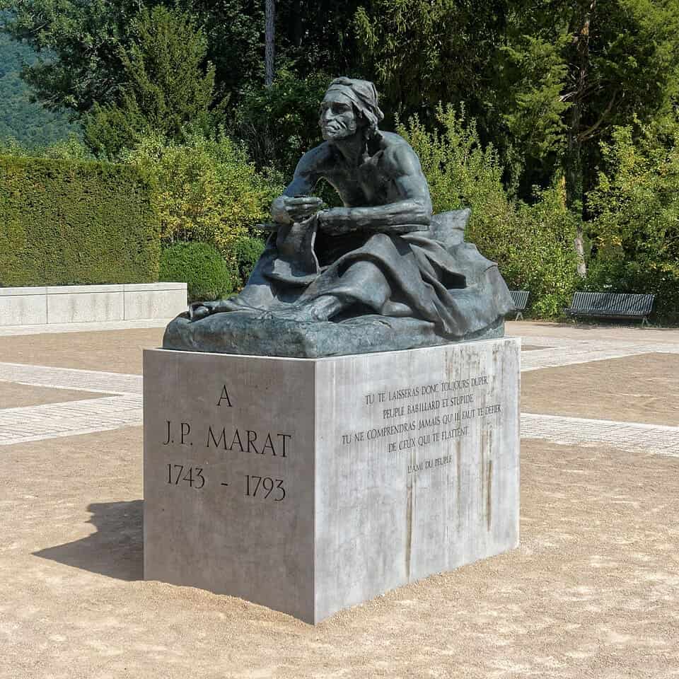 Escultura en bronce de Jean-Paul Marat sentado sobre un pedestal de piedra con inscripciones conmemorativas.