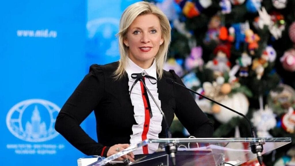 María Zakharova en atril oficial con chaqueta negra y blusa blanca con volantes sobre fondo azul del Ministerio de Asuntos Exteriores de Rusia.