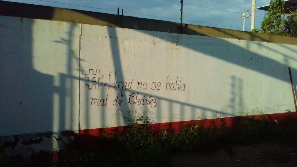 Muro pintado de propaganda chavista con la frase escrita en rojo Aquí no se habla mal de Chávez junto a etiqueta estilizada.