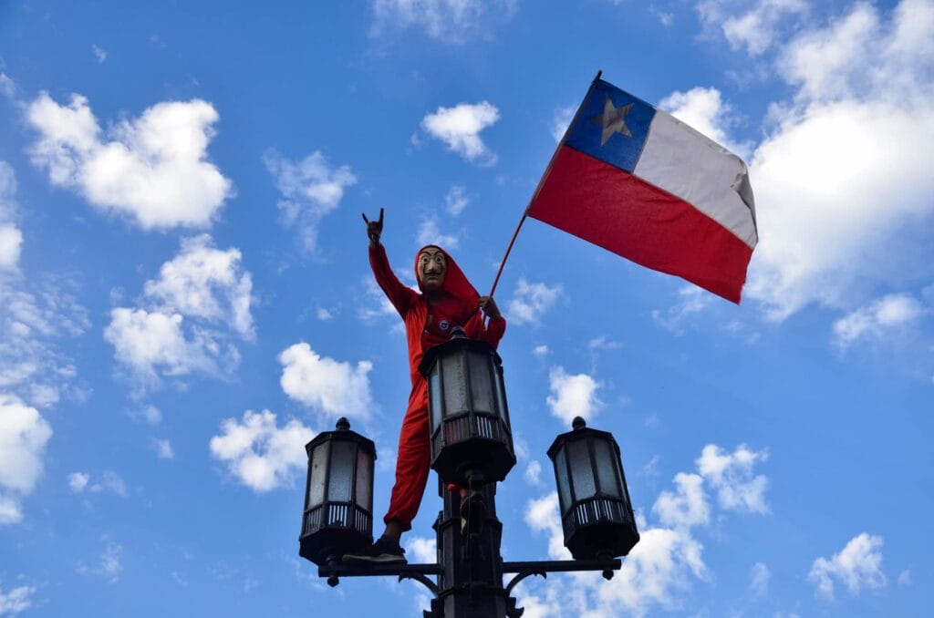 Manifestante disfrazado con mono rojo y máscara de Dalí sobre un farol ondeando la bandera de Chile bajo un cielo azul con nubes.