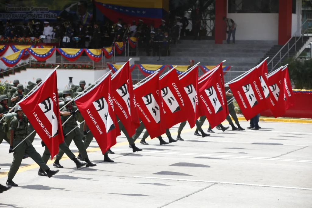 Soldados desfilando con banderas rojas de propaganda chavista con la frase Chávez vive la lucha sigue durante un acto militar.